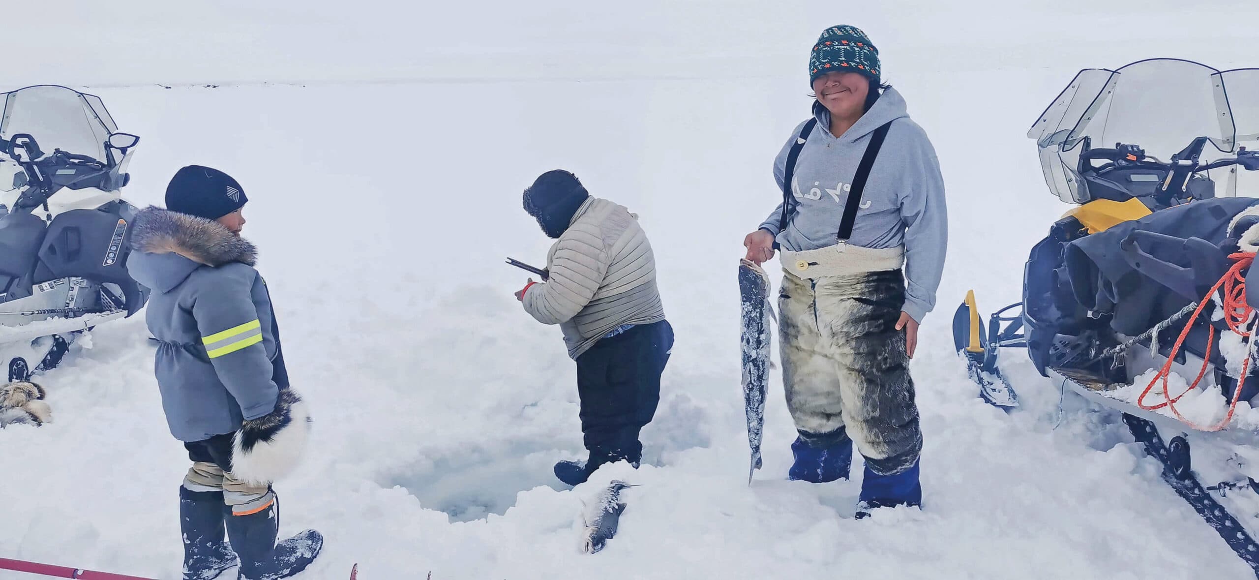 Three people ice fishing near Pond Inlet, Canada. One. person is holding a fish. There are two snowmobiles in the background.