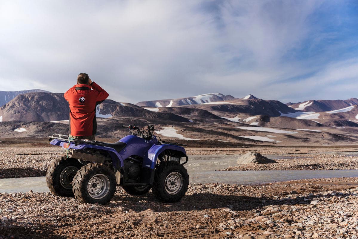 Man standing by a ATV with binoculars over looking a snowy and muddy landscape.