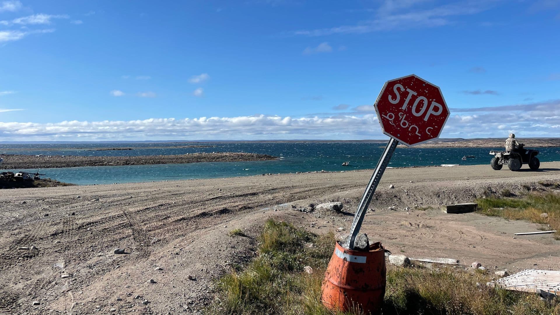 Stop sign written in English letters and Inuktitut syllabics. The sign is bent to the side and the coast is in the. background.