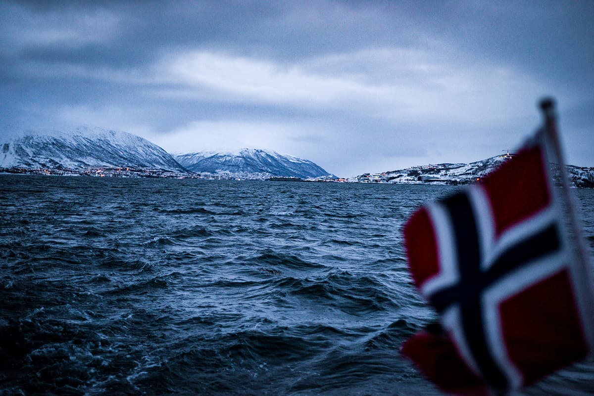 Norwegian flag in the foreground. Snowy mountains and a town in the background.
