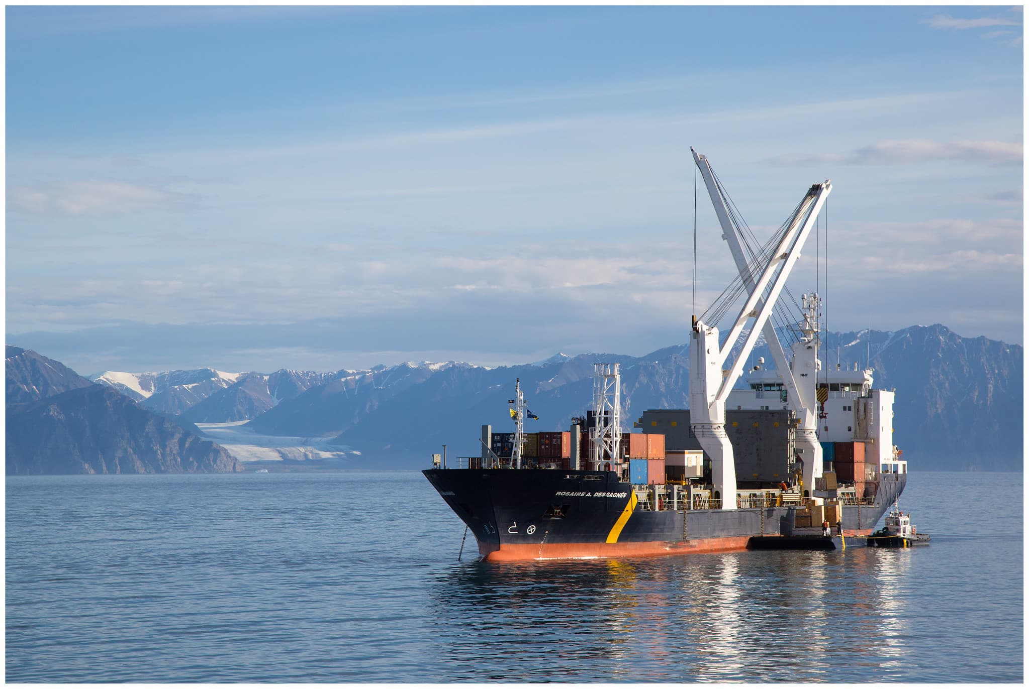 Large ship with shipping containers surrounded by snow-capped mountains in the distance.